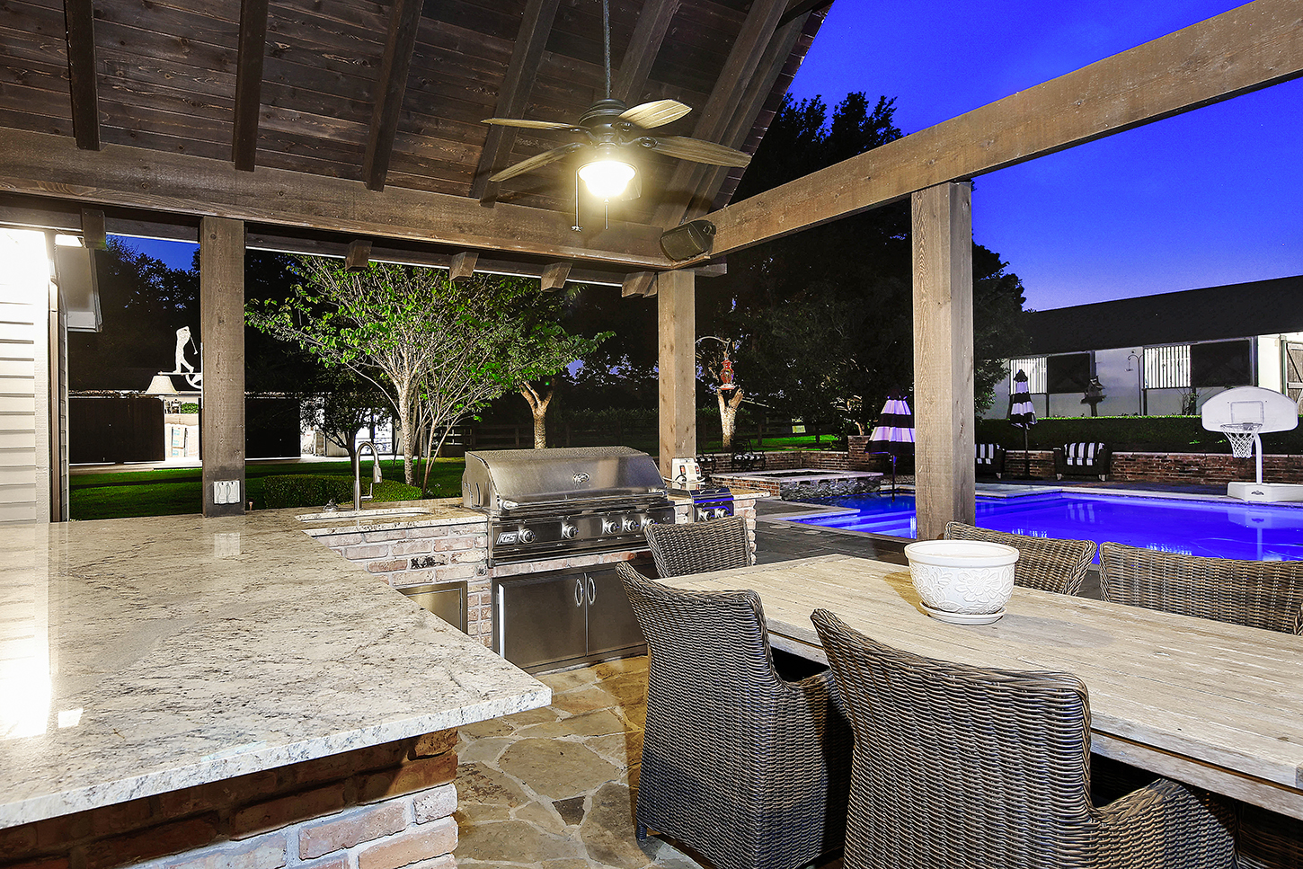 Covered outdoor kitchen with grill and dining beside the pool at twilight